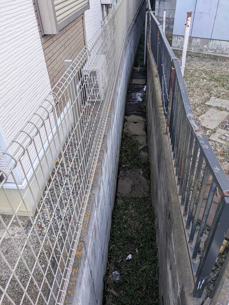 既存の傾いた擁壁と歪んだフェンス・フェンス横の水路